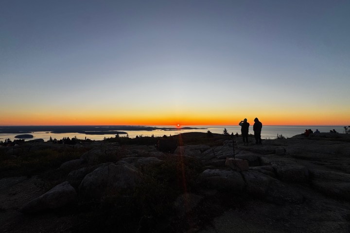 Two people silhouetted against a sunrise on a rocky hill overlooking water.