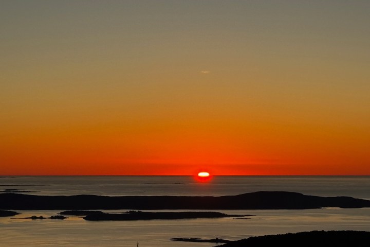 Sunset over ocean, horizon with silhouetted islands and vivid orange sky.