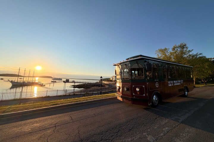 Red trolley near a coastal sunset with boats and calm water.
