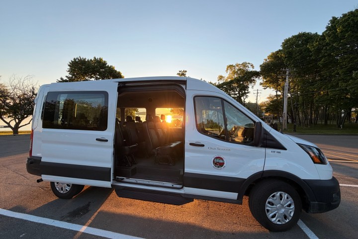 White van with open side door, sunset reflecting inside, parked in empty lot with trees.