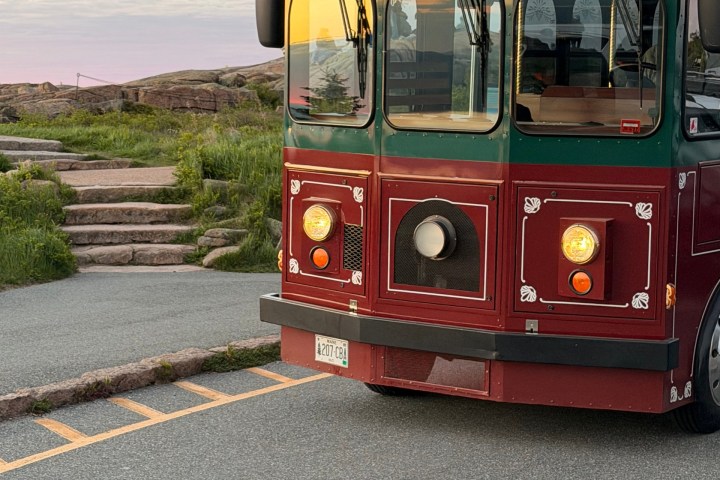 Red and green trolley parked on roadside near stone steps and grass.